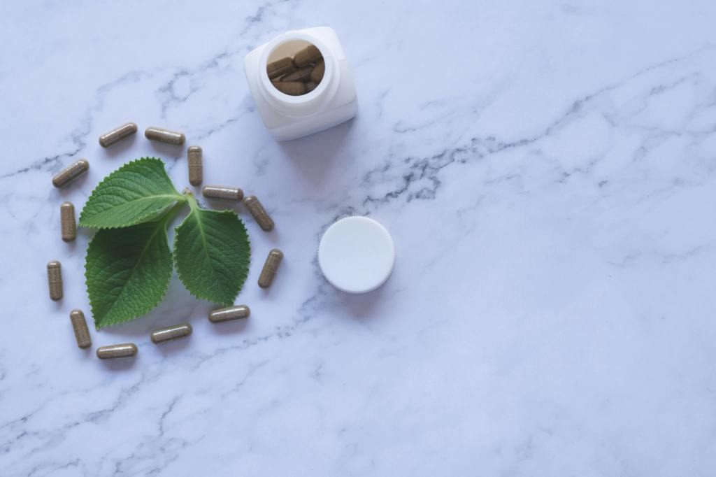 Herbal supplement capsules arranged in a circle around fresh green leaves, with an open white bottle and its cap on a marble surface. Caption below reads '- Chitrak: The One That Made Me Sweat (Literally).