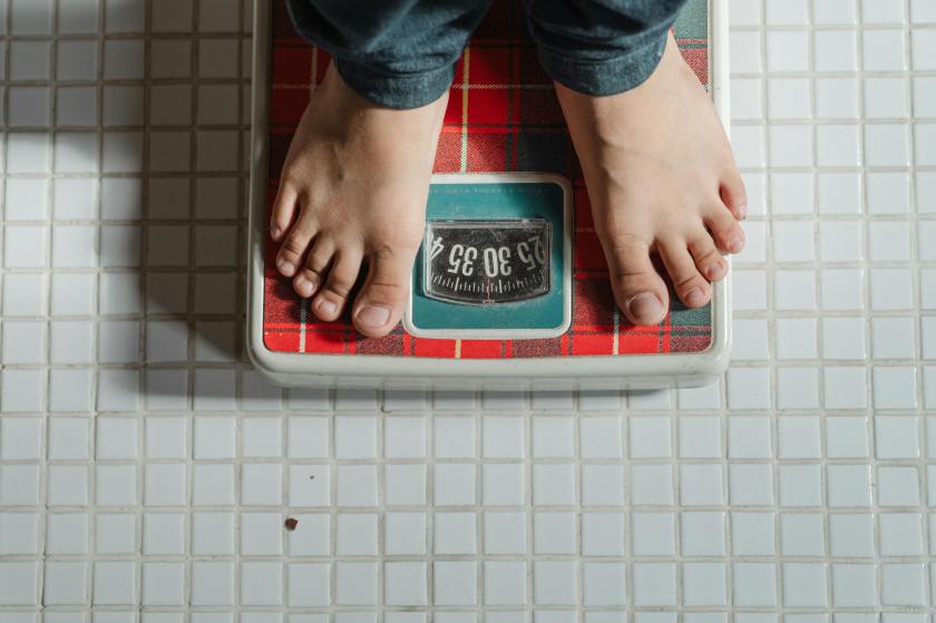 Overhead view of bare feet standing on a vintage red plaid bathroom scale on white tiled floor, representing weight management challenges. Appears alongside the quote 'Control what you can, when you can, and forgive yourself for the rest.